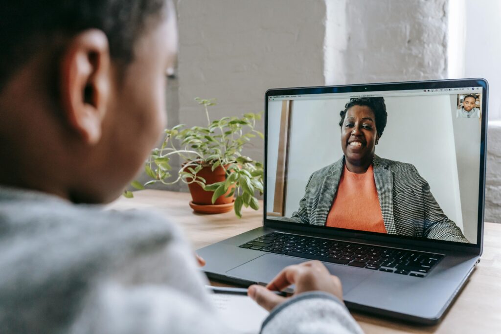 Crop anonymous African American kid sitting at table with modern laptop and listening to female teacher explaining lesson during coronavirus pandemic