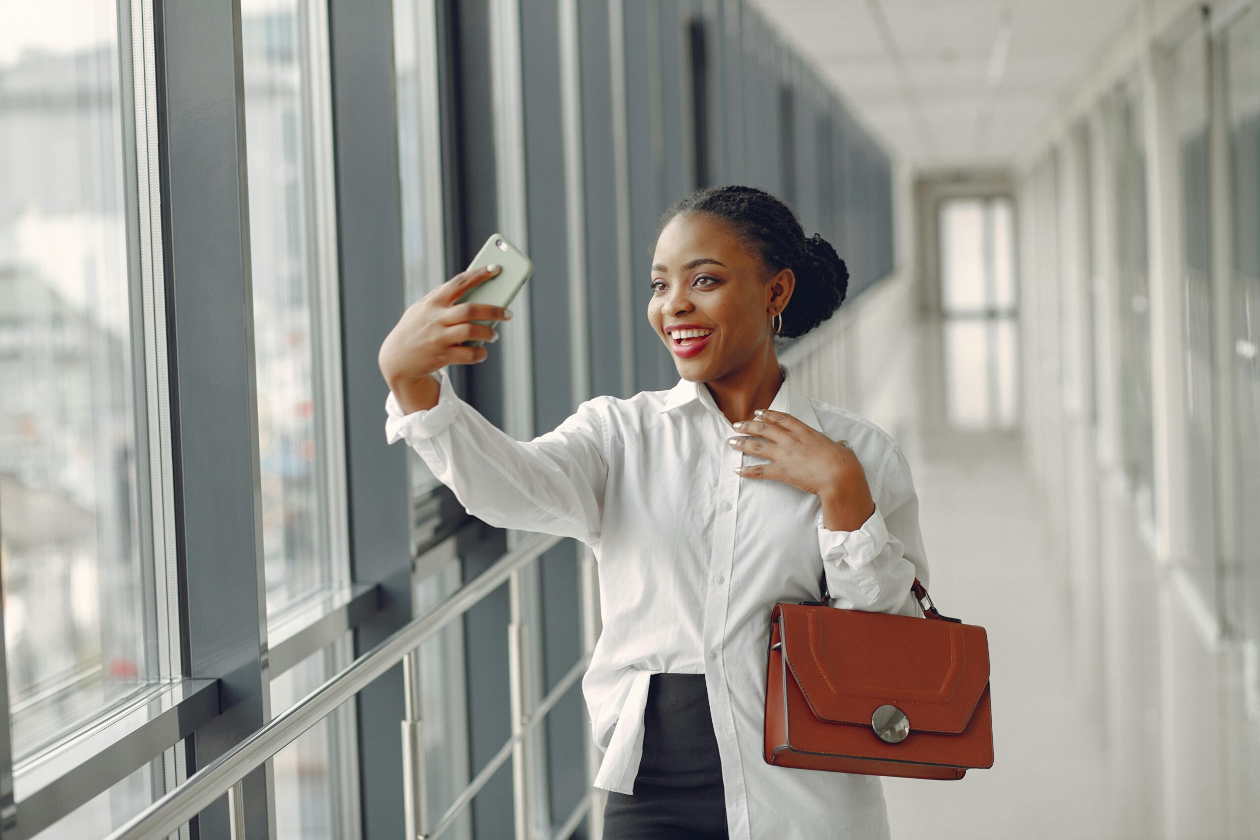 Cheerful young black female office worker in formal wear with stylish handbag taking selfie while walking along glass corridor in contemporary office building
